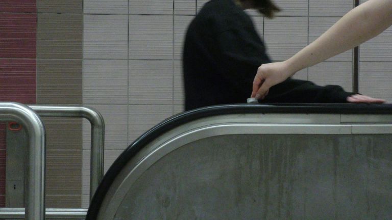 Eine Hand, die mit einem Eiswürfel auf dem Handlauf einer Rolltreppe entlangfährt