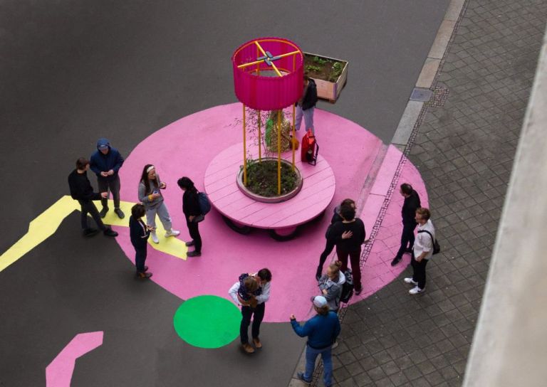Von oben fotografierte Szene auf der Straße mit mehreren Menschen, die sich um eine pinke runde Bank und einen pinken auf der Straße aufgemalten Punkt versammeln