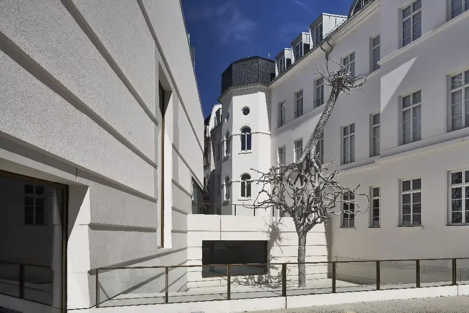 Skulptur mit zwei an den Kronen ineinanderverschlungene Bäume, bei denen einer in der Erde verwurzelt ist, während der Stamm und die Wurzeln des anderen in den Himmel ragen auf Museumsvorplatz des Jüdischen Museums, Eingang des Neubaus links, rechts Teile des Altbaus